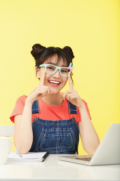 Smiling Asian girl with topknots, in brightly colored glasses sitting at desk with laptop