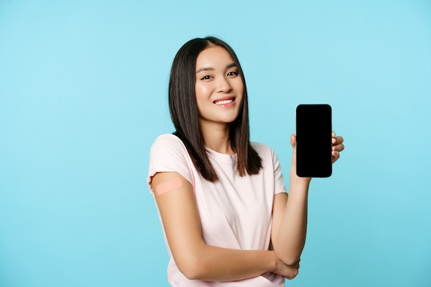 백신을 접종한 팔을 들고 웃고 있는 아시아 소녀, 휴대폰 빈 화면, 건강 여권 개념, 스마트폰에 covid-19 예방 접종 증명서