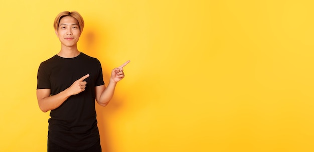 Free photo smiling asian male student with blond hair pointing fingers right showing logo standing yellow backg