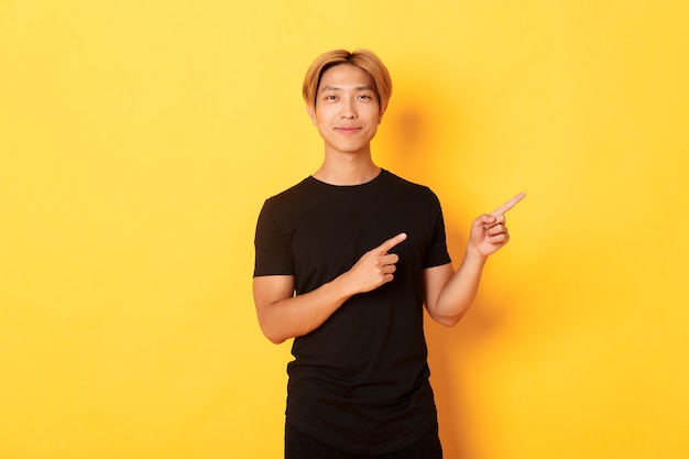 Smiling asian male student with blond hair, pointing fingers right, showing logo, standing yellow wall.