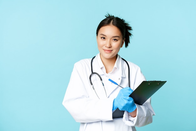 Smiling asian medical worker female physician writing down patient info holding pen and clipboard st...