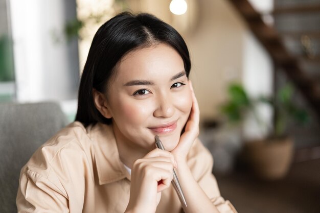 Smiling asian millennial girl working from home holding pen and looking cute at camera doing homewor...