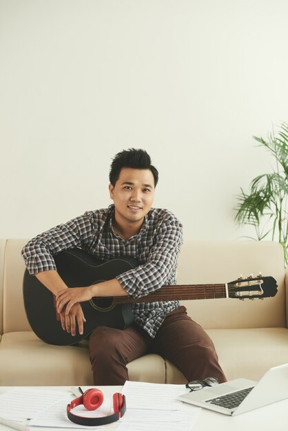 Smiling Asian musician sitting on couch with guitar at home