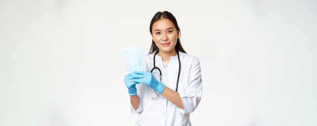 Улыбающийся азиатский врач семейный врач показывает стерильные маски для лица, предотвращающие заражение covid19, стоя в униформе на белом фоне