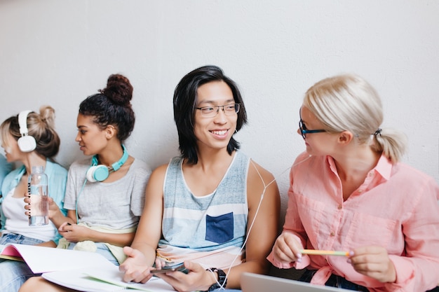 Smiling asian student discussing favorite song with blonde woman during preparing lessons. Indoor portrait of pleased university friends talking about exams amd listening music in white headphones.