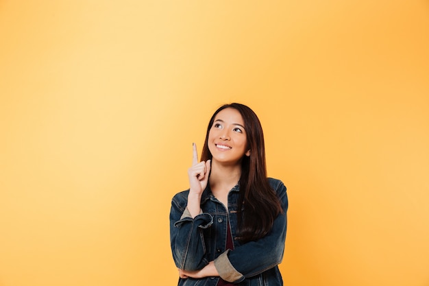 Smiling asian woman in denim jacket pointing and looking up over yellow background