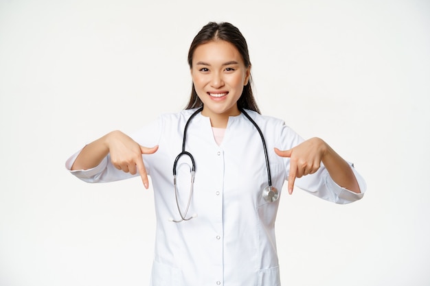 Smiling asian woman doctor pointing fingers down and showing promo deal discounts on clinic service ...
