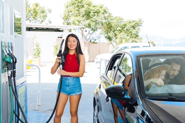 Smiling Asian woman holding fuel pump nozzle and looking at camera while friends exploring map 