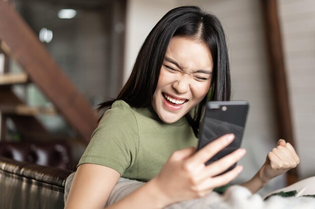 웃고 있는 아시아 여성이 만족스럽게 주먹을 쥔 채 집에 앉아 휴대폰을 읽고 있습니다.