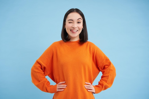 Smiling asian woman looks confident winks with enthusiastic face expression stands over blue studio