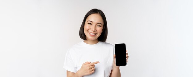 Smiling asian woman pointing finger at smartphone screen showing application interface mobile phone website standing over white background