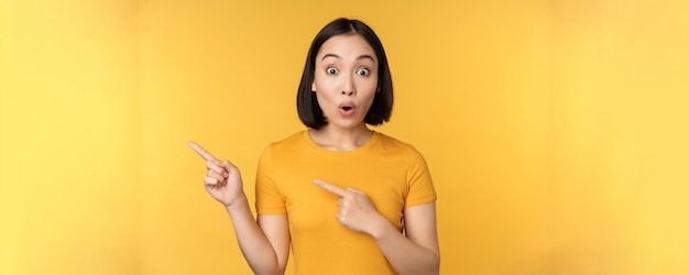 Smiling asian woman pointing fingers left showing advertisement on empty copy space standing over yellow background