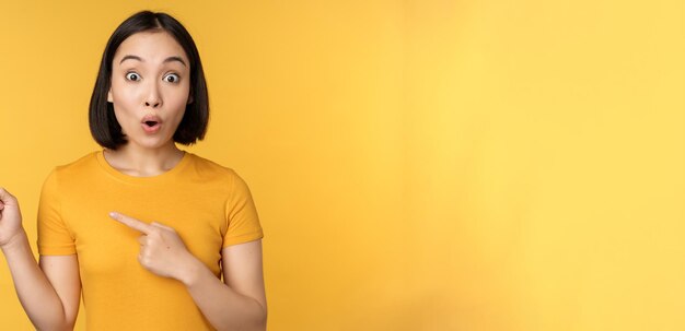 Smiling asian woman pointing fingers left showing advertisement on empty copy space standing over yellow background