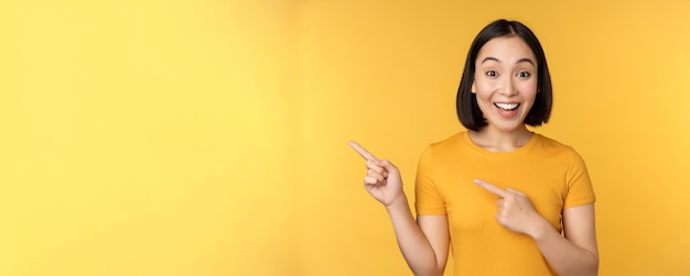 무료 사진 노란색 배경 위에 서 있는 빈 카피 공간에 광고를 보여주는 왼쪽 손가락을 가리키는 웃는 아시아 여성
