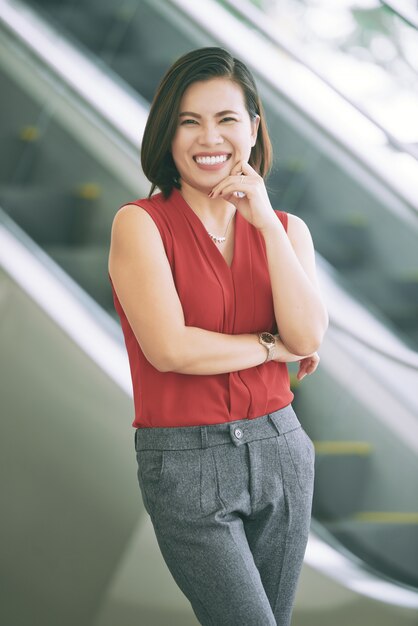 Smiling Asian woman posing in front of escalator with finger touching cheek