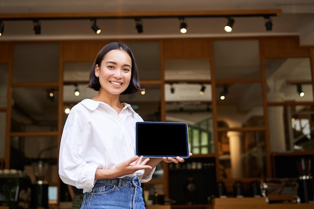 Улыбающаяся азиатка показывает владельца кафе на экране цифрового планшета, показывающего что-то стоящее перед кафе