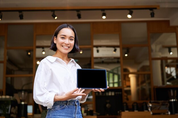 カフェの前に立ってスマートを示すデジタル タブレット スクリーン カフェ オーナーを示すアジアの女性の笑みを浮かべてください。