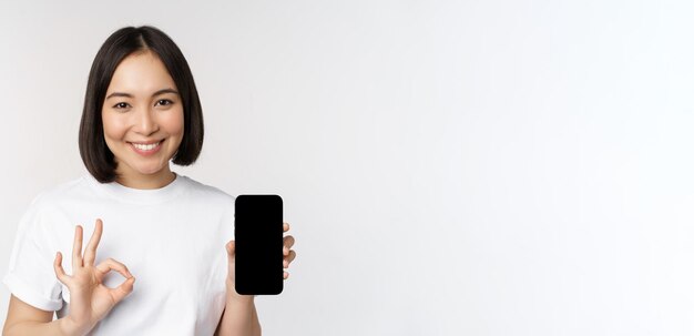 Smiling asian woman showing okay mobile phone screen recommending smartphone app standing over white background