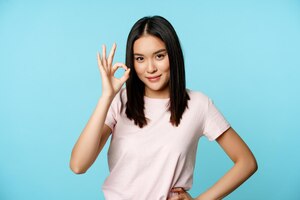 Улыбающаяся азиатская женщина, показывающая хороший знак, дает одобрение, рекомендует что-л. с хорошей репутацией на синем фоне