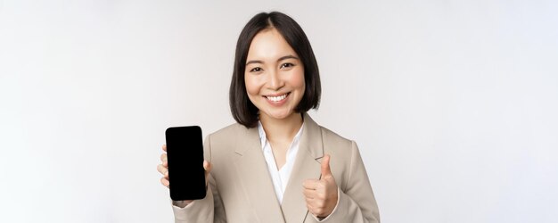 Smiling asian woman showing smartphone screen and thumbs up Corporate person demonstrates mobile phone app interface standing over white background