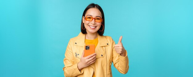 Smiling asian woman showing thumbs up recording on mobile phone using smartphone app and recommending it standing over blue background