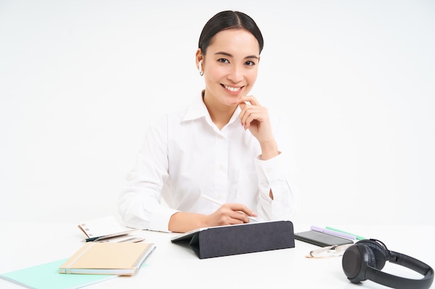 Free photo smiling asian woman sits in her office works in digital tablet has headphones smartphone and work do