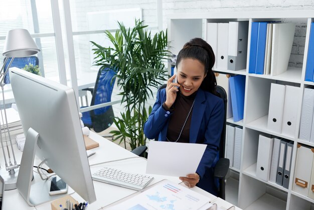 Smiling Asian woman sitting at desk in office, looking at document and talking on mobile phone