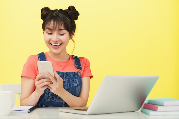 Donna asiatica sorridente che si siede allo scrittorio con il computer portatile e i libri e che per mezzo dello smartphone