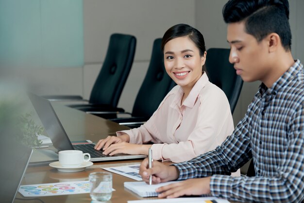 Smiling Asian woman sitting at meting table with laptop, amd male colleague writing in notebook