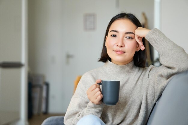 家でコーヒーを飲み、仕事の後にくつろぐマグカップでソファに座っている笑顔のアジア人女性