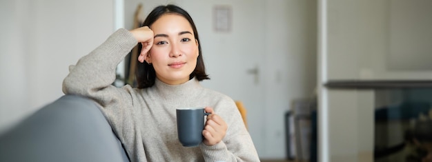 집에서 커피를 마시고 퇴근 후 휴식을 취하며 머그잔을 들고 소파에 앉아 웃고 있는 아시아 여성