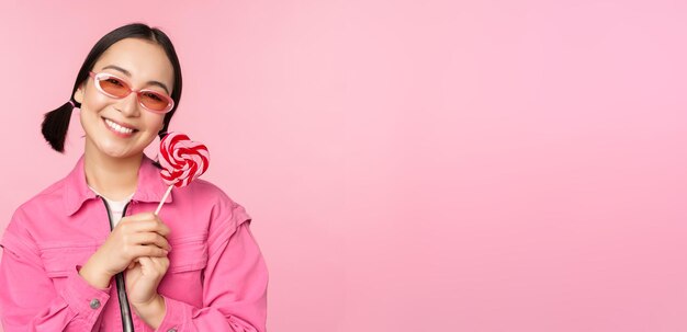 Smiling asian woman in sunglasses holding lolipop sweets eating candy and looking happy standing over pink background