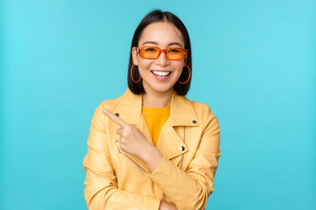 Smiling asian woman in sunglasses pointing finger left showing banner logo or advertisement standing over blue background Copy space