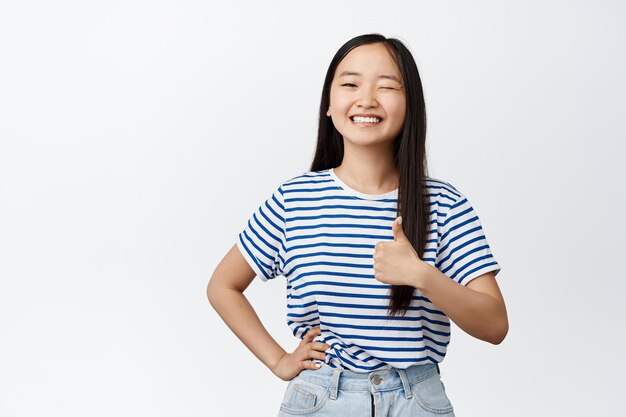 Smiling asian woman winking at camera showing thumb up in approval encourage you praise good work standing satisfied against white background