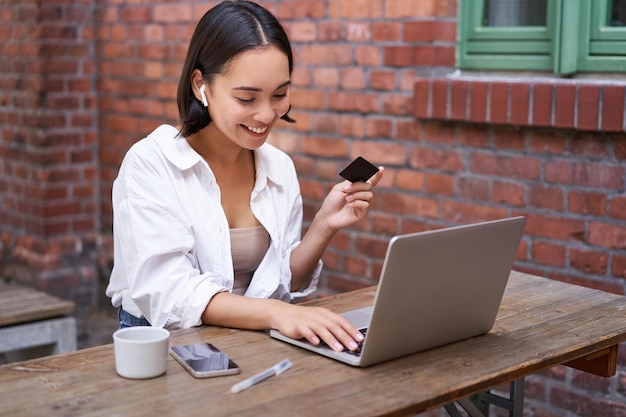 Smiling asian woman with laptop and wireless earphones paying with credit card buying online sitting