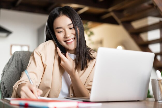 Smiling asian woman working from home taking notes in notebook talking on cellphone and writing down...