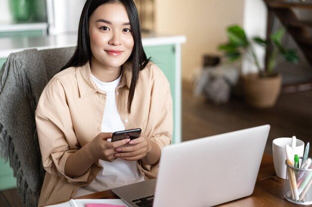 컴퓨터 근처에 앉아 스마트폰을 들고 집에서 노트북으로 일하는 웃는 아시아 여성