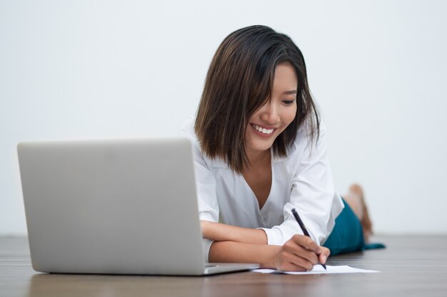 Smiling Asian Woman Writing on Floor with Laptop