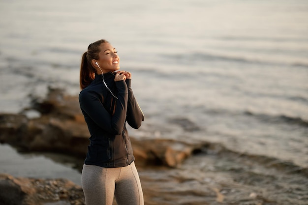 Smiling athletic woman standing at coastline with her eyes closed while enjoying in music over earphones Copy space