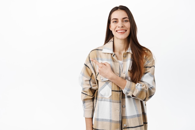 Smiling attractive girl demonstrate promo deal, showing copyspace aside, pointing finger left at advertisement and looking pleased, recommending, standing over white background