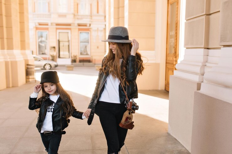 Free Photo smiling attractive girl in felt hat with retro camera walking down the street holding little sister's hand. magnificent young mother in leather jacket with daughter searching nice place for photoshoot