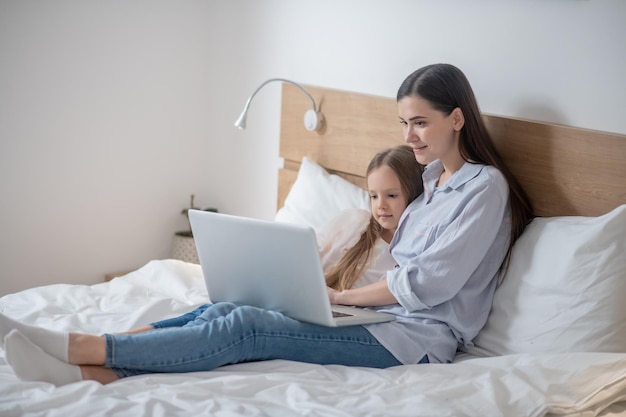 Улыбающаяся привлекательная женщина и ее милый спокойный ребенок сидят на кровати перед ноутбуком