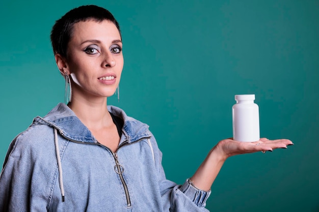 Smiling attractive woman holding bottle of pills presenting health care product at camera while standing over isolated background in studio. Cheerful female reviewing pharmaceutical supplement