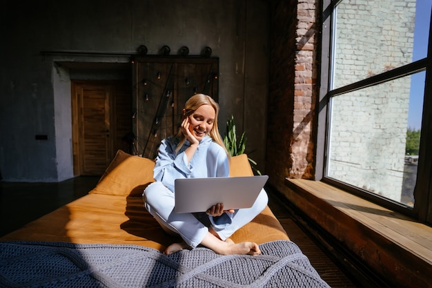 Donna attraente sorridente che si trova sul letto facendo uso del computer portatile