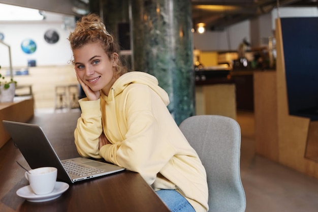 커피를 마시고 노트북을 사용하는 카페에 앉아 웃는 매력적인 여자 카페에서 원격 작업하면서 행복 카메라를보고 아름다운 소녀 노트북을 사용하여 야외에서 공부하는 여학생