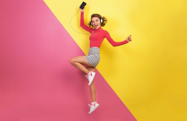 Smiling attractive woman in stylish colorful outfit jumping and listening to music in headphones