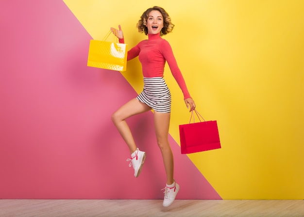 Smiling attractive woman in stylish colorful outfit jumping with shopping bags, happy, pink yellow background, polo neck, striped mini skirt, sale, discout, shopaholic, fashion summer trend, emotional