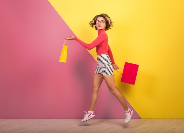 Smiling attractive woman in stylish colorful outfit jumping with shopping bags on pink yellow background, polo neck, striped mini skirt, shopaholic on sale, fashion summer trend