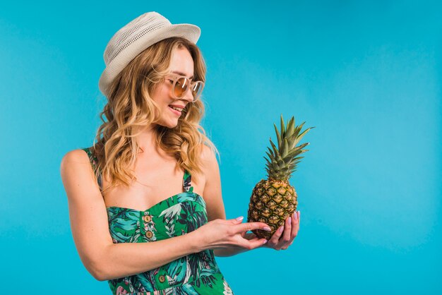 Улыбаясь привлекательная молодая женщина в платье с шляпу и солнцезащитные очки, глядя на свежий ананас