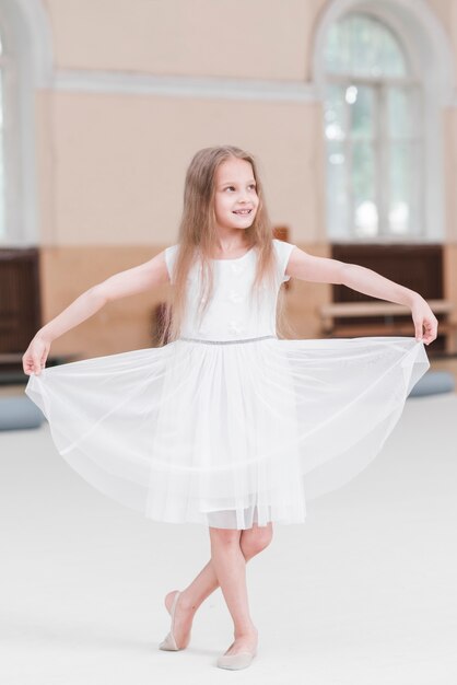 Smiling ballerina girl with crossed leg holding white her dress